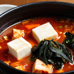 A steaming pot of Kimchi stew (Kimchi-jjigae) with pork belly, tofu, and green onions, garnished with 김가루 (seaweed flakes). Focus on the rich, red broth and appetizing presentation.